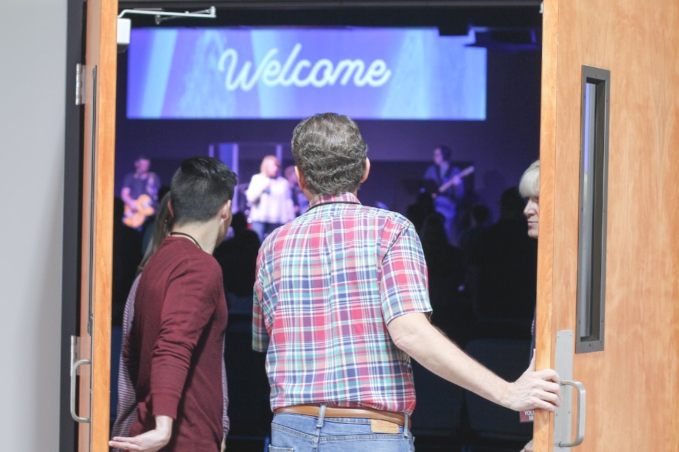 Men entering doors of Worship Center with band on stage and Welcome sign at back.