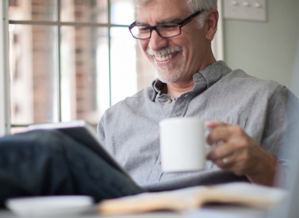 Older smiling man holding coffee while viewing tablet with open Bible at forefront.