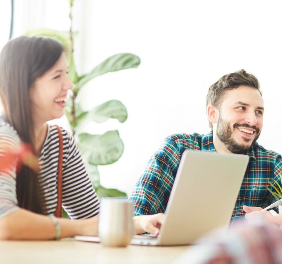 Smiling woman and man looking right with laptop at desk with mug.